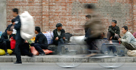 A CYCLIST RIDES PAST MIGRANT WORKERS AT A LONG DISTANCE BUS STATION IN BEIJING.